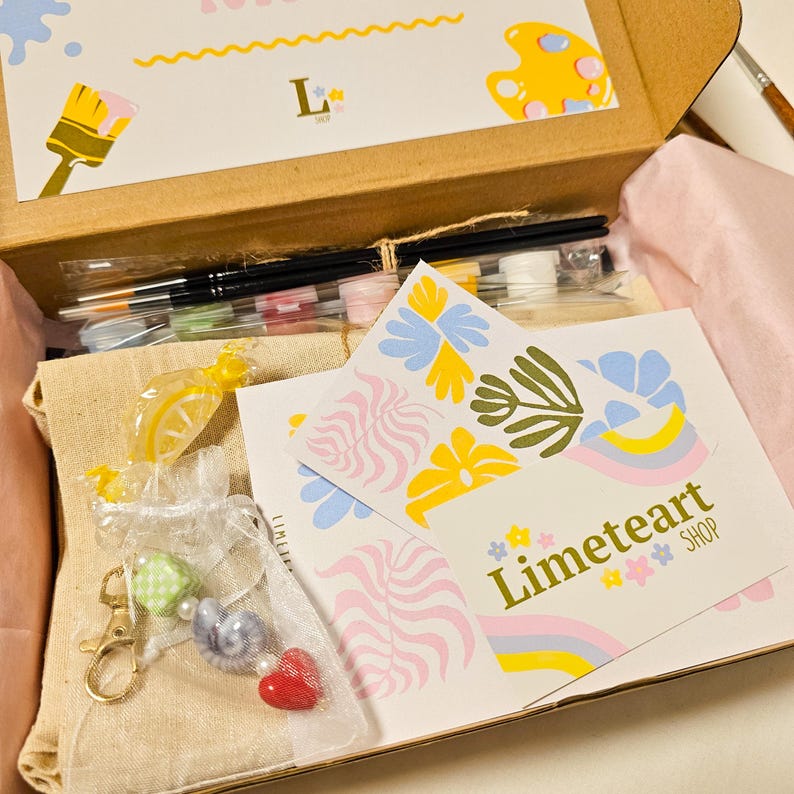 May include: An open cardboard box filled with art supplies. Includes paintbrushes, paint, and decorative items. The box contains a card with the text "Limeteart SHOP" and floral designs. A small bag with a heart charm is visible.
