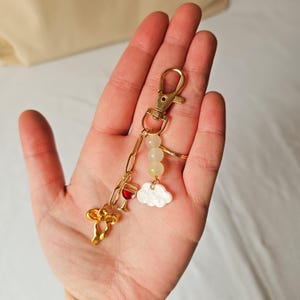 Puede incluir: Llavero dorado con un dije de copa de vino, un dije de nube y cuentas blancas. El llavero est&aacute; unido a un anillo de metal dorado.