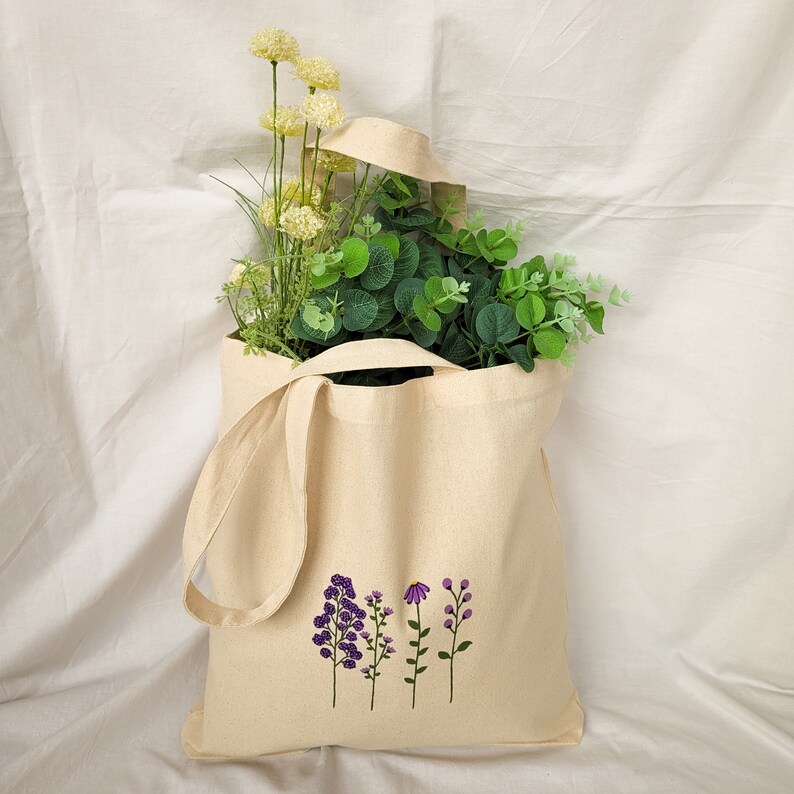 May include: A white canvas tote bag with a design of purple flowers on the front. The bag has long handles and is filled with green and white flowers.