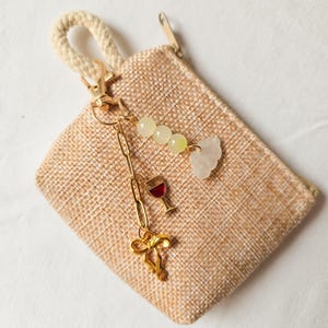 Puede incluir: Una peque&ntilde;a bolsa de arpillera beige con un tirador de cremallera dorado. Una cadena dorada con abalorios cuelga del tirador de la cremallera, incluyendo un abalorio de copa de vino, un abalorio de nube y varios abalorios de cuentas verdes y blancas.