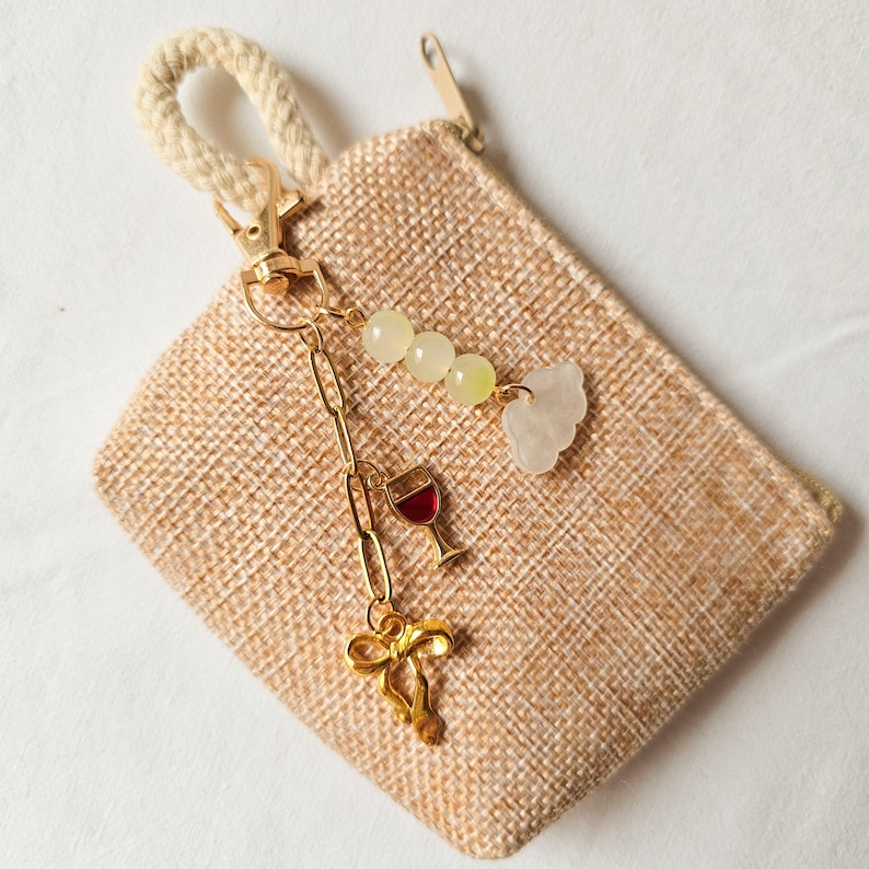 Puede incluir: Una peque&ntilde;a bolsa de arpillera beige con un tirador de cremallera dorado. Una cadena dorada con abalorios cuelga del tirador de la cremallera, incluyendo un abalorio de copa de vino, un abalorio de nube y varios abalorios de cuentas verdes y blancas.