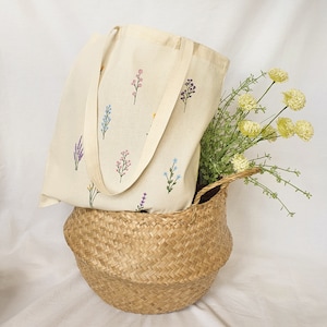May include: A white canvas tote bag with embroidered floral designs in shades of blue, pink, and yellow. The bag is sitting in a woven wicker basket.