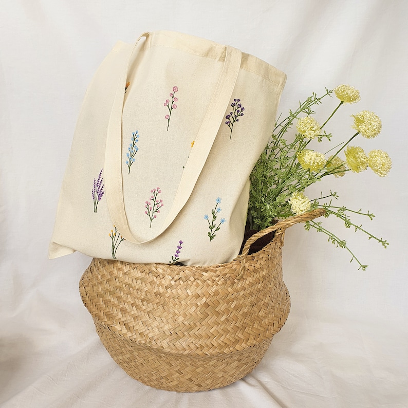May include: A white canvas tote bag with embroidered floral designs in shades of blue, pink, and yellow. The bag is sitting in a woven wicker basket.