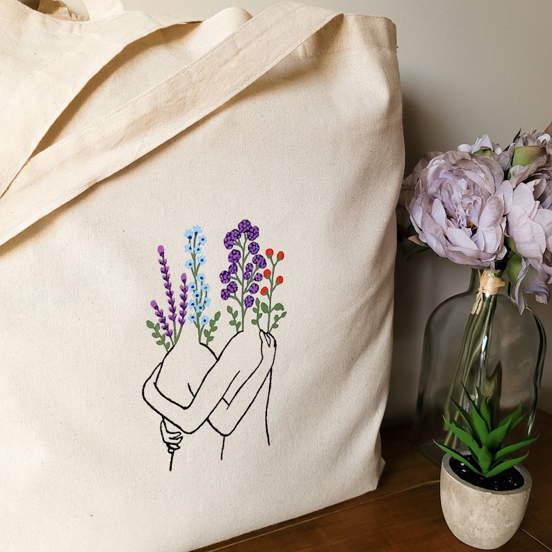 May include: A white tote bag with an embroidered design of two figures embracing, each with a bouquet of purple, blue, and red flowers.