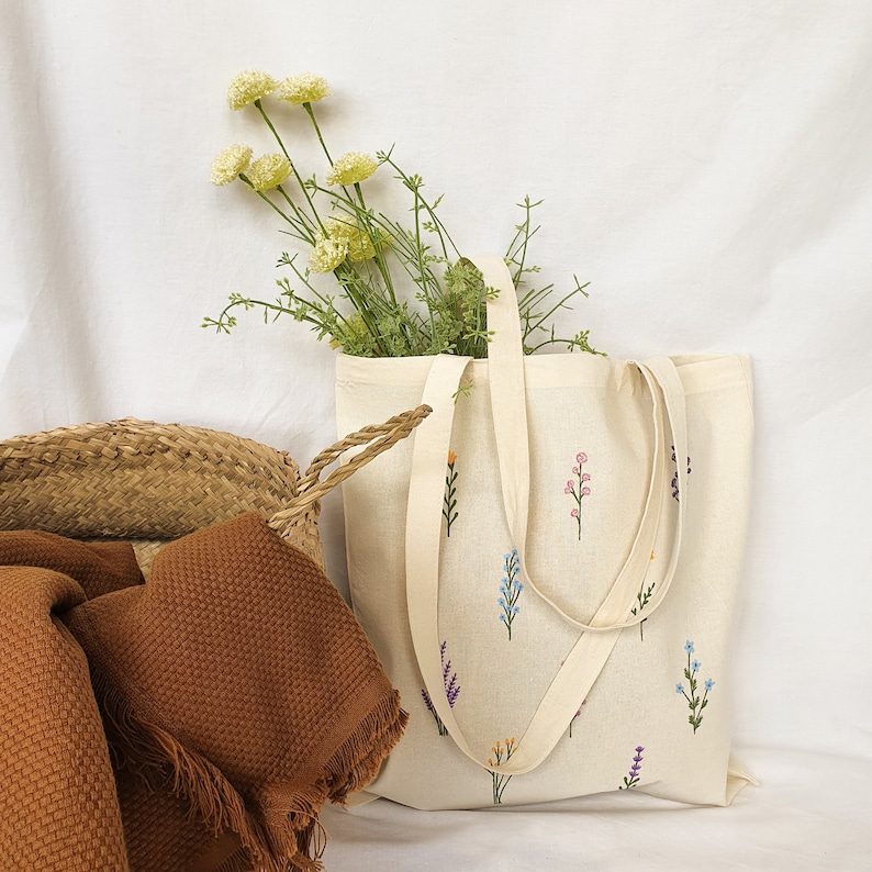 May include: A white canvas tote bag with embroidered floral designs in shades of pink, blue, and green. The bag has two handles and a wide base.