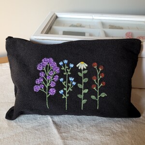 May include: Black canvas zippered pouch with embroidered flowers in shades of purple, blue, white, green, and red.