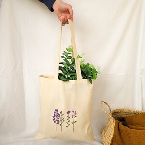 May include: A beige tote bag with a design of four purple flowers on the front. The bag is made of canvas and has a long handle.