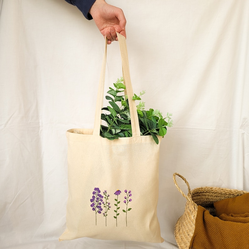 May include: A beige tote bag with a design of four purple flowers on the front. The bag is made of canvas and has a long handle.