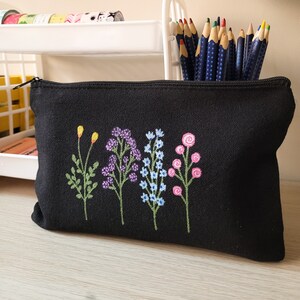 May include: Black canvas zippered pouch with embroidered flowers in shades of pink, blue, purple, and yellow. The flowers are stylized and have a whimsical feel.