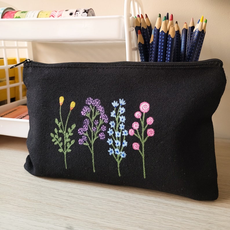 May include: Black canvas zippered pouch with embroidered flowers in shades of pink, blue, purple, and yellow. The flowers are stylized and have a whimsical feel.