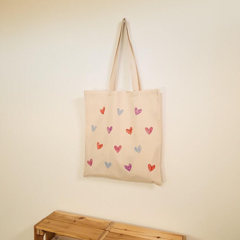 May include: A beige canvas tote bag with a pattern of colorful hearts. The hearts are in shades of pink, red, blue, and purple.