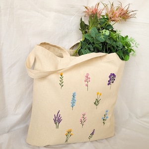 May include: A beige tote bag with embroidered flowers in various colors, including blue, yellow, pink, and purple. The bag has two handles and is made of a natural fabric.