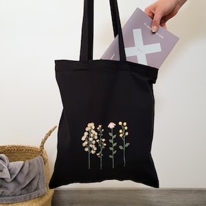 May include: Black tote bag with embroidered flowers in shades of pink, yellow, and green. The bag has a book tucked inside with the text "el secreto a vivir" and "know" visible.
