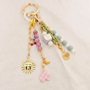 Może przedstawiać: Złoty brelok do kluczy z wieloma zawieszkami. Zawieszki obejmują słońce, wiśnie, księżyc, różowe serce w szachownicę oraz sznury koralików w kolorze różowym, zielonym i niebieskim. Brelok ma złoty pierścień i zapięcie.
