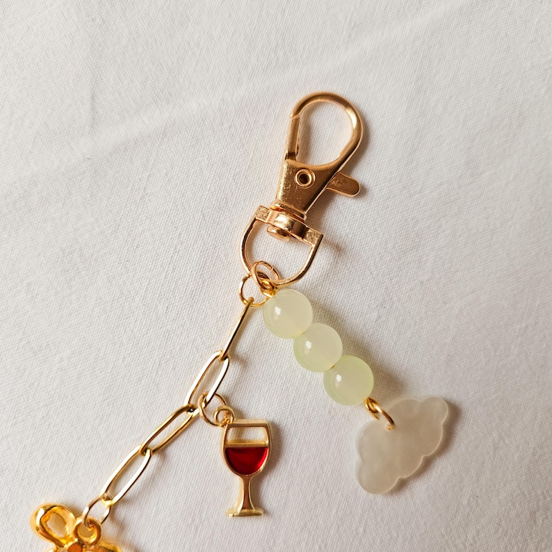 Puede incluir: Llavero dorado con un dije de copa de vino tinto, un dije de nube blanca y una cadena de cuentas verde claro.
