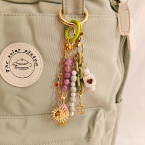 May include: A collection of colorful charms and beads hang from a gold-colored keychain. The charms include a gold sun, pink leaf, and various beads in shades of purple, green, and white. The keychain is attached to a light green backpack.
