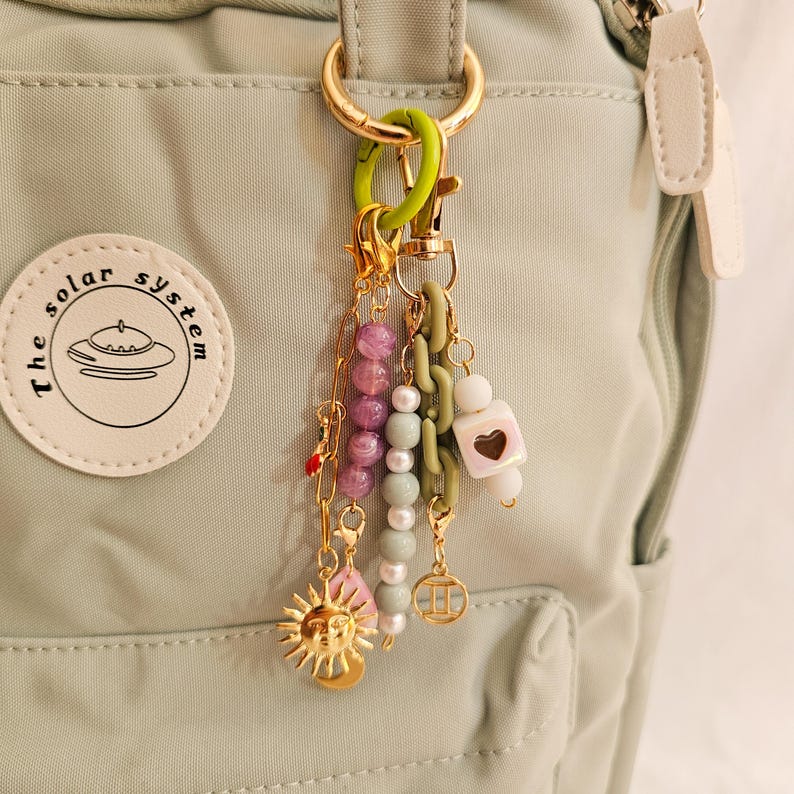 May include: A collection of colorful charms and beads hang from a gold-colored keychain. The charms include a gold sun, pink leaf, and various beads in shades of purple, green, and white. The keychain is attached to a light green backpack.