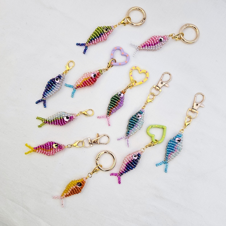 May include: A collection of beaded fish keychains in various colors, including pink, blue, green, and yellow. Each keychain features a gold-colored clasp and ring. The fish have detailed beadwork and are arranged on a white surface.