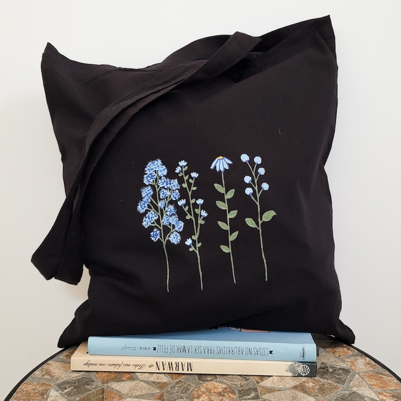 May include: Black tote bag with a floral design embroidered on the front. The flowers are blue and green and are arranged in a vertical line. The bag is sitting on top of a book with the title "COSAS NO ACONTECEM PARA SER FELIZ" and the author "MARWAN".