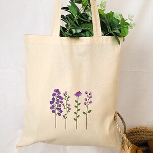 May include: A white canvas tote bag with a design of purple flowers on the front. The flowers are stylized and have green stems.