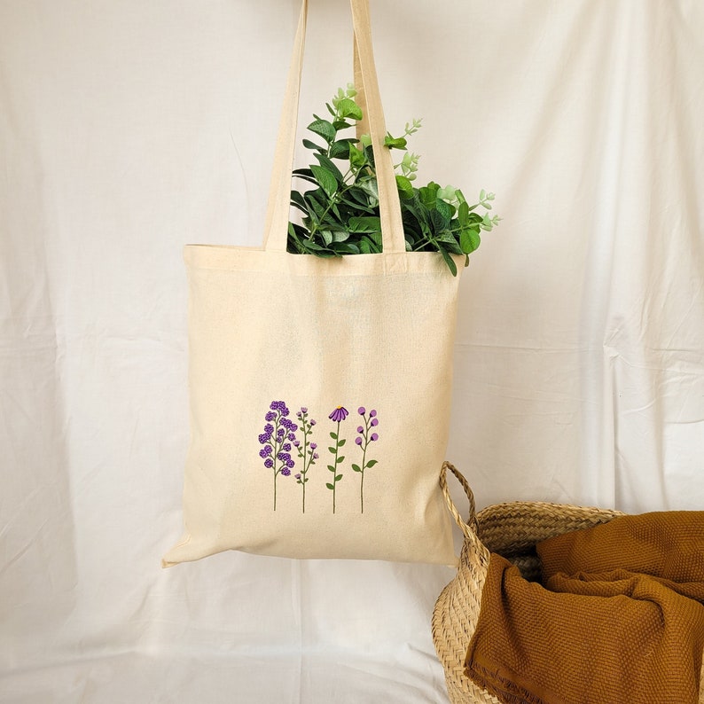 May include: A beige tote bag with a floral design featuring purple and pink flowers. The bag has a long handle and is made of canvas.