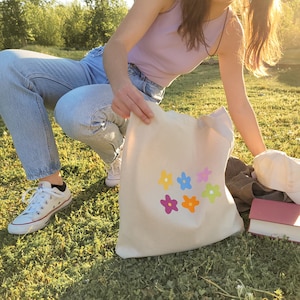 Peut inclure: Un sac fourre-tout en toile blanche avec des motifs de fleurs multicolores. Le sac est tenu par une personne portant un jean bleu et un d&eacute;bardeur violet clair.