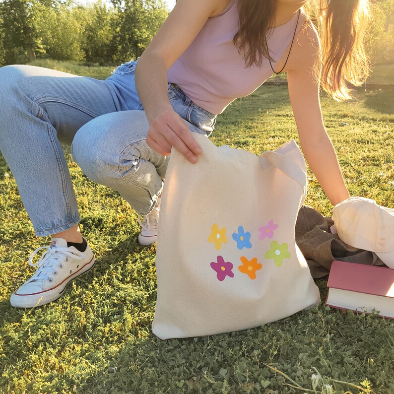 Peut inclure: Un sac fourre-tout en toile blanche avec des motifs de fleurs multicolores. Le sac est tenu par une personne portant un jean bleu et un d&eacute;bardeur violet clair.