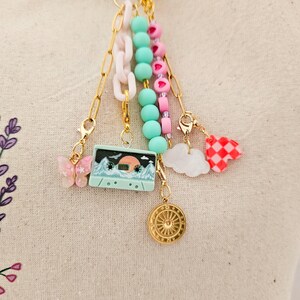 May include: A collection of colorful charms and beads on gold chains. Charms include a pink butterfly, a cassette tape with a mountain scene, a gold medallion, a white cloud, and a red and white checkered heart.