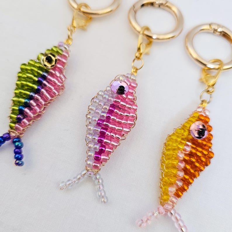May include: Three beaded fish keychains in vibrant colors. One is green, pink, and blue; another is pink and white; and the third is yellow and orange. Each fish has a gold-colored clasp and a beaded tail, perfect for attaching to keys or bags.