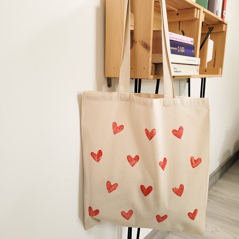 May include: A beige canvas tote bag with red heart designs printed on the front. The bag has two handles and is hanging on a wooden shelf.