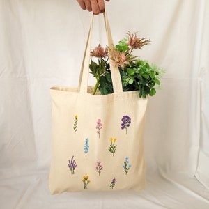 May include: A beige tote bag with embroidered flowers in various colors, including purple, pink, blue, and yellow. The bag has a long handle and is made of a natural fabric.