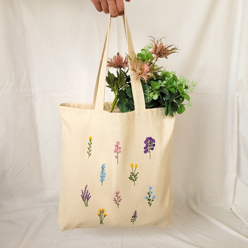 May include: A beige tote bag with embroidered flowers in various colors, including purple, pink, blue, and yellow. The bag has a long handle and is made of a natural fabric.