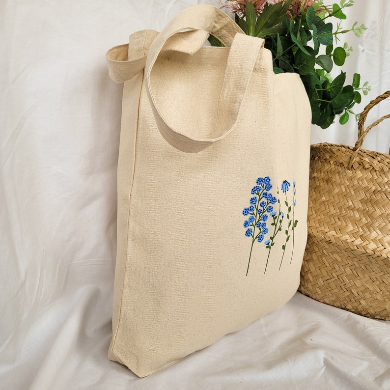 Peut inclure: Un sac fourre-tout en toile beige avec une longue bandouli&egrave;re. Le sac est d&eacute;cor&eacute; d'un motif de fleurs bleues brod&eacute;es sur le devant.