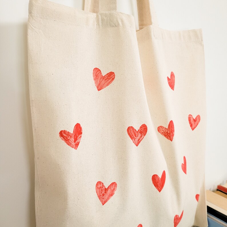 May include: A white canvas tote bag with red heart designs printed on it. The bag has two handles.