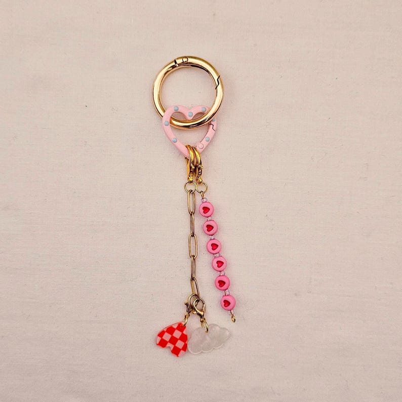 May include: A gold-colored keychain with a pink heart-shaped charm featuring blue polka dots. The keychain has a chain with pink heart beads and charms of a red and white checkered shape and a white cloud.