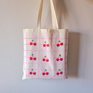 May include: A white canvas tote bag with pink grid pattern and red cherries printed on it.