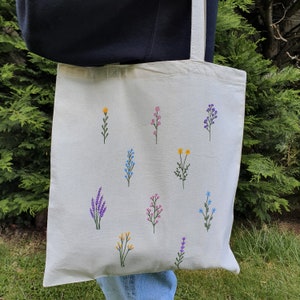 May include: A white canvas tote bag with a variety of embroidered flowers in different colors, including yellow, pink, blue, and purple.