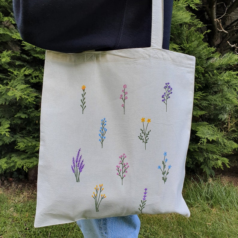 May include: A white canvas tote bag with a variety of embroidered flowers in different colors, including yellow, pink, blue, and purple.