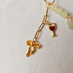 Puede incluir: Cadena de oro con un dije de lazo de oro, un dije de copa de vino de oro con esmalte rojo y cuentas de jade verde.