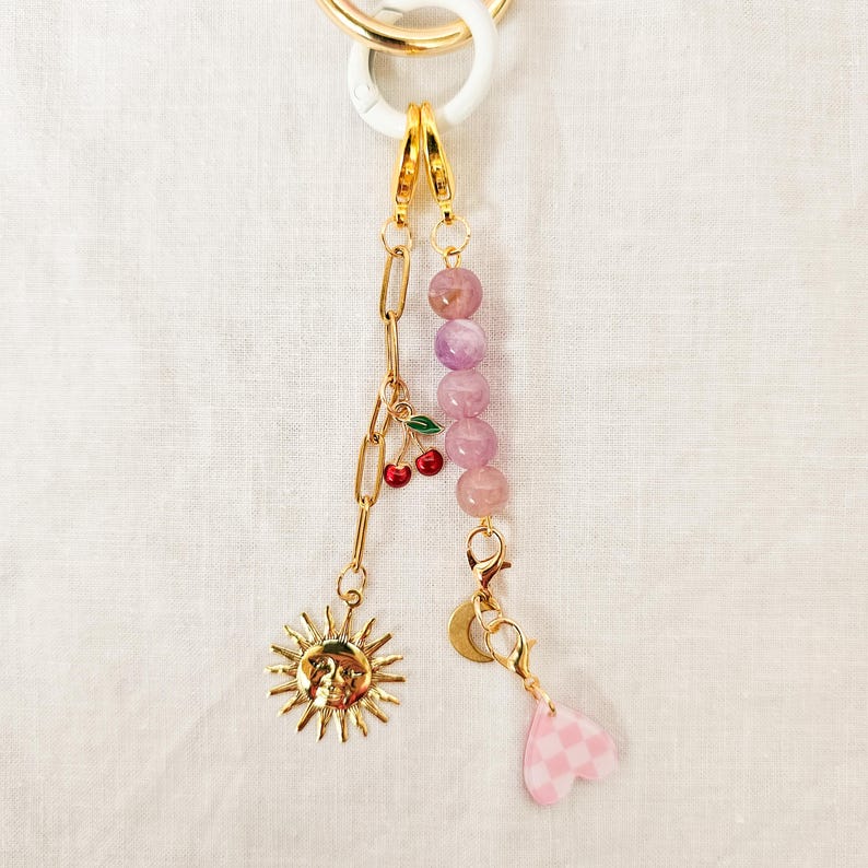 May include: A gold-toned keychain with a white ring, featuring a chain, sun, cherry, and pink heart charm. The chain has a cherry charm with red cherries and green leaves. The keychain also has a strand of pink beads.