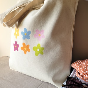 Peut inclure: Un sac fourre-tout en toile beige avec un motif de fleurs color&eacute;es. Les fleurs sont dans des tons de rose, jaune, orange et vert.
