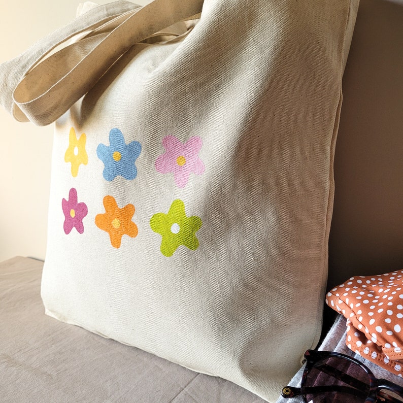 Peut inclure: Un sac fourre-tout en toile beige avec un motif de fleurs color&eacute;es. Les fleurs sont dans des tons de rose, jaune, orange et vert.