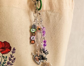 May include: A silver keychain with a green clasp, adorned with charms. The charms include colorful Halloween-themed beads, a cross, a heart, a spider, and a spiderweb. The keychain is attached to a beige tote bag with a floral design.