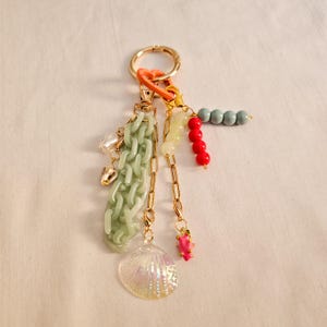 以下が含まれることがあります： さまざまなチャームが付いたゴールドトーンのキーホルダー。チャームには、ライトグリーンのチェーン、貝殻、パール、赤、黄、青のカラフルなビーズが含まれます。オレンジ色の留め金がキーホルダーをゴールドリングに接続します。