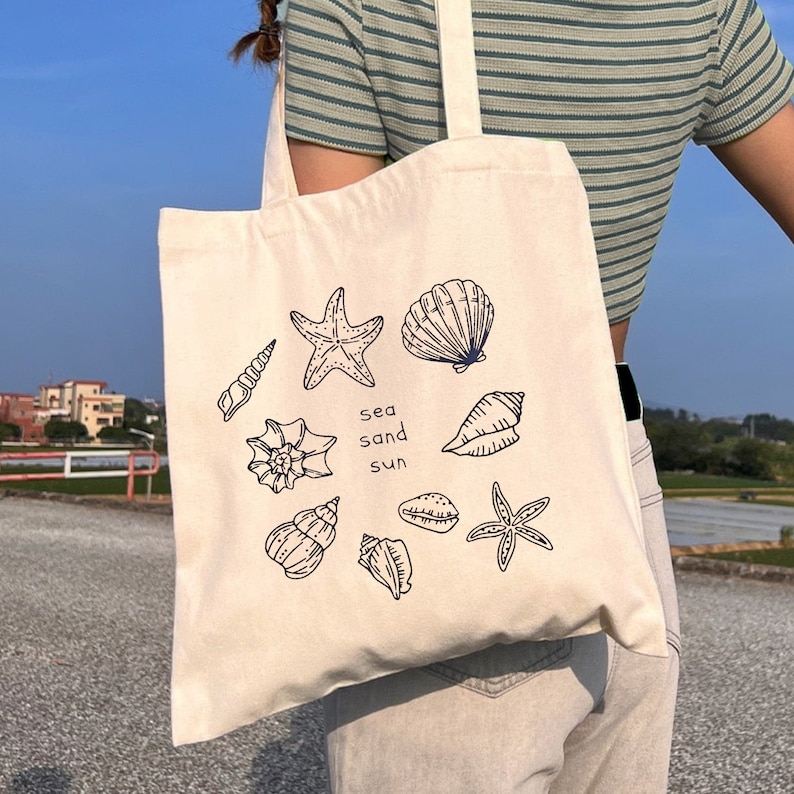 May include: A white canvas tote bag featuring a black and blue design of seashells, starfish, and the words "sea sand sun".