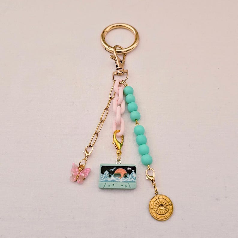 May include: A gold-toned keychain featuring three dangling charms: a pink butterfly, a light blue cassette tape with a mountain and sun graphic, and a round gold medallion with a sunburst design, accented by pink links and turquoise beads.