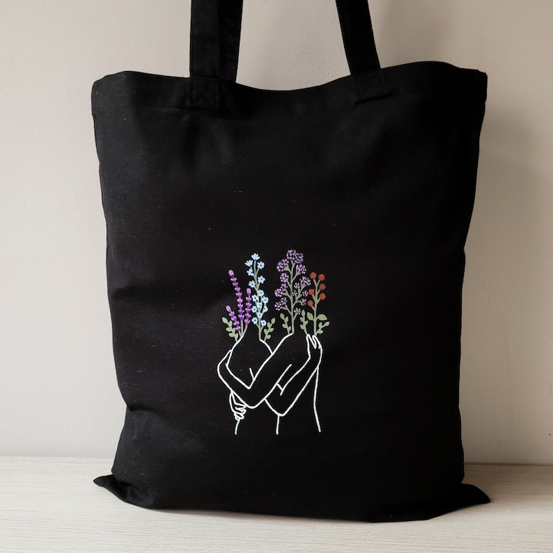 May include: Black tote bag with a white line drawing of two figures embracing. The figures have flowers growing from their heads, including purple, blue, and red.
