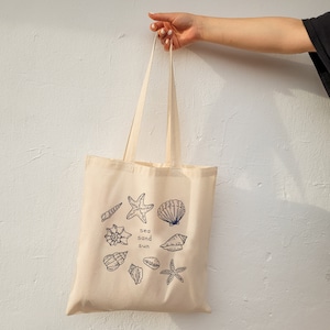 May include: A beige canvas tote bag with a blue design of seashells, starfish, and the words "sea sand sun".