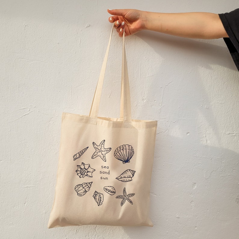 May include: A beige canvas tote bag with a blue design of seashells, starfish, and the words "sea sand sun".