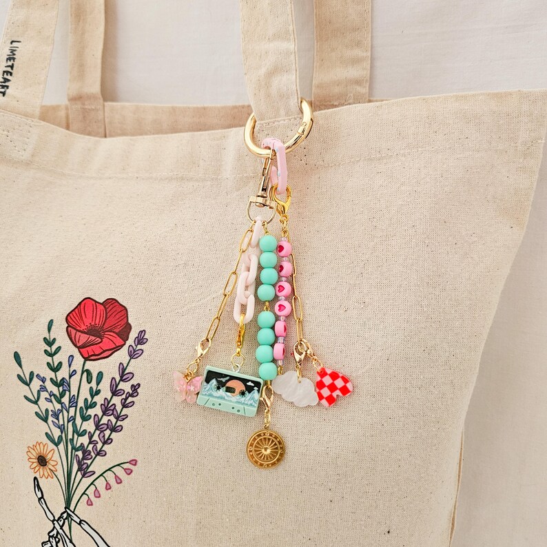 May include: A gold-toned keychain with various charms, including a cassette tape, a butterfly, and a compass. The keychain also features pink, mint green, and white beads, and a red and white checkered heart charm. The keychain is attached to a beige tote bag.